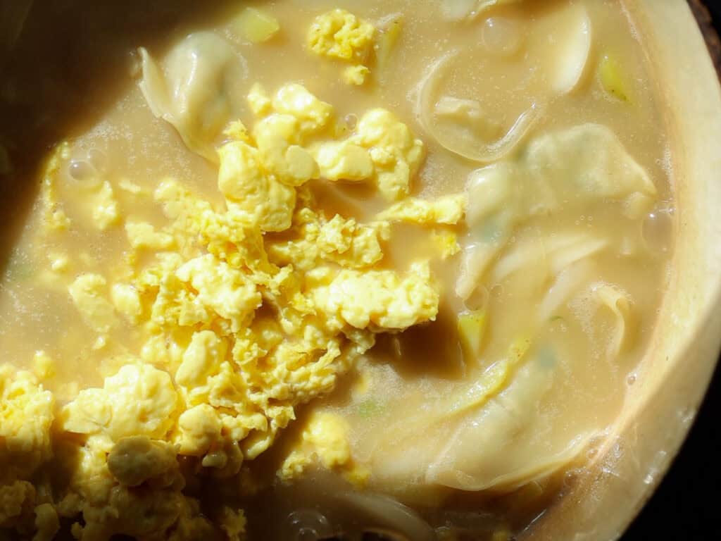 A close-up of a bowl of creamy vegan rice cake soup containing scrambled vegan eggs, dumplings, and sliced onions. The broth appears rich and thick, with a golden hue and pieces of cooked egg floating on the surface.